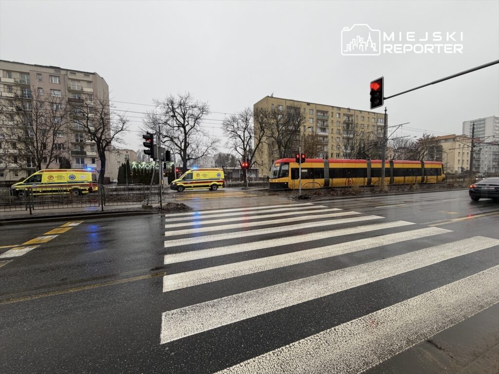 Dwa karetki pogotowia z włączonymi sygnałami świetlnymi zatrzymane na skrzyżowaniu, obok tramwaj wstrzymany na czerwonym świetle.
