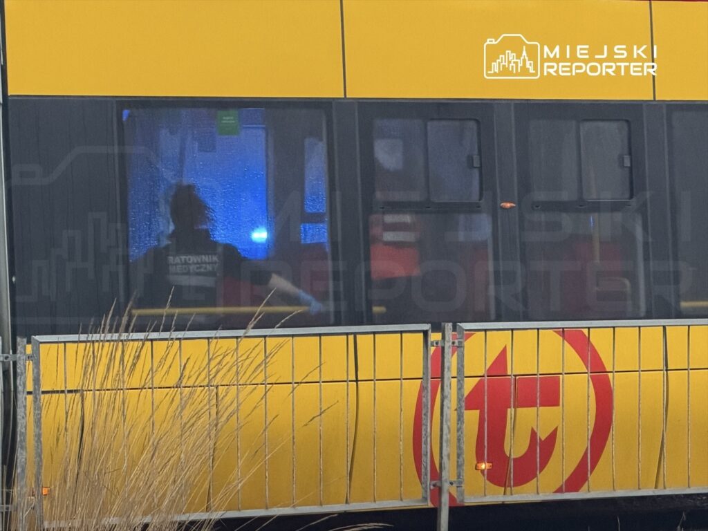 Ratownik medyczny w samochodzie pogotowia, z niebieskimi światłami w tle, w miejskim otoczeniu.