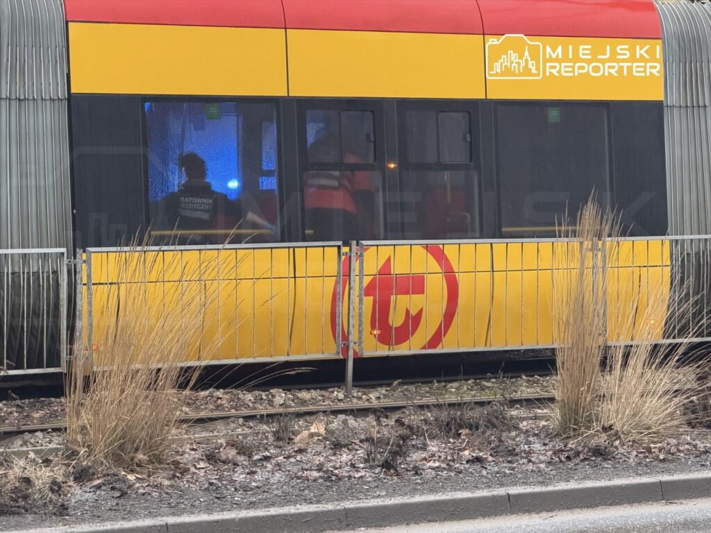 Ratownik medyczny w czerwonej kurtce udziela pomocy w tramwaju, w tle widać migające niebieskie światła.
