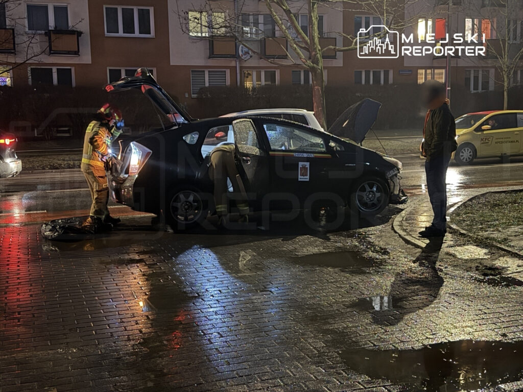 Strażak z narzędziami otwiera bagażnik samochodu osobowego na oświetlonej ulicy, obok stoi mężczyzna.