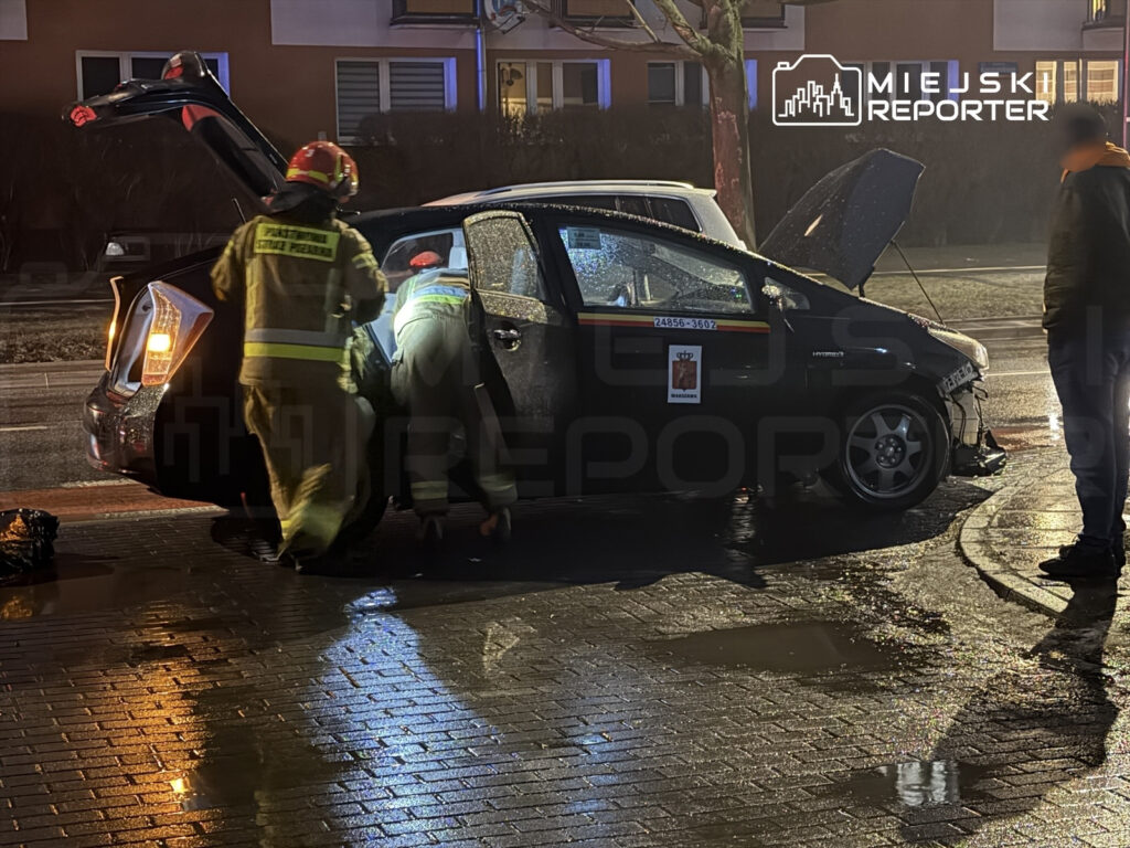 Strażacy sprawdzają wnętrze uszkodzonego samochodu zaparkowanego na ulicy, w tle widoczne oświetlenie nocne.