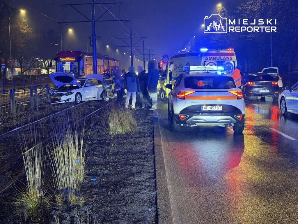 W nocy na ulicy, zderzenie dwóch samochodów; z białego pojazdu unosi się dym, a ratownicy udzielają pomocy poszkodowanym.