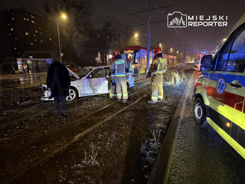 Kierowca w białym samochodzie osobowym stoi obok pojazdu uszkodzonego na torach tramwajowych, obok interweniują strażacy.
