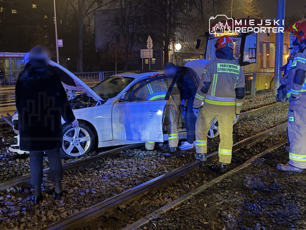 Mężczyzna w czarnym płaszczu obserwuje działania strażaków przy unieruchomionym samochodzie na torach tramwajowych.