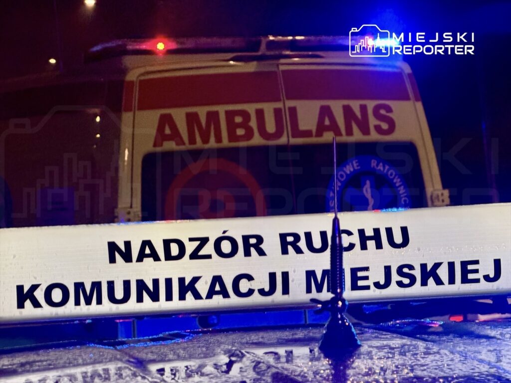 Ambulans z włączonymi sygnalizatorami stojący obok znaku "Nadzór Ruchu Komunikacji Miejskiej" w nocy.
