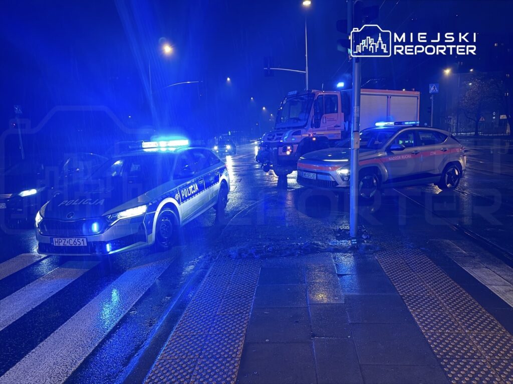 Radiowóz policyjny z włączonymi sygnałami świetlnymi blokuje skrzyżowanie w deszczowy wieczór. Obok stoi pojazd nadzoru ruchu.