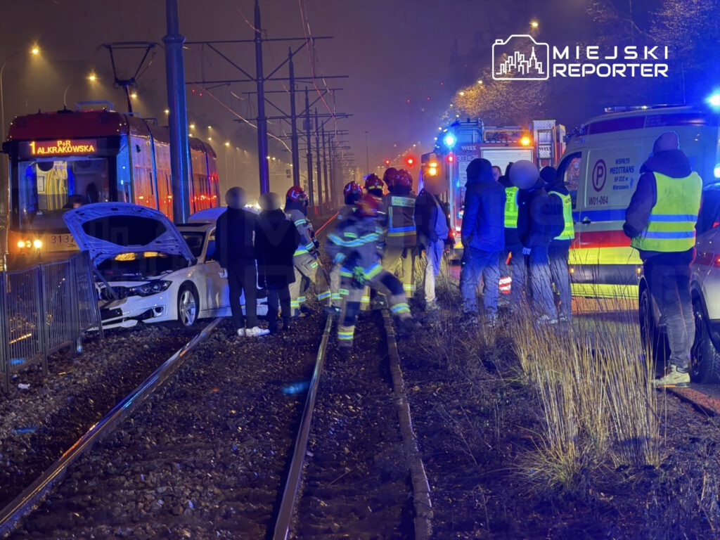 Grupa ratowników i policjantów pracuje przy uszkodzonym samochodzie na torach tramwajowych w nocy.