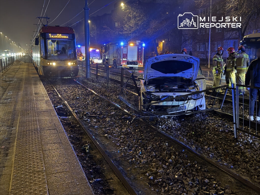 Tramwaj zatrzymany na torach, obok widać uszkodzony samochód osobowy oraz grupę strażaków w akcji.