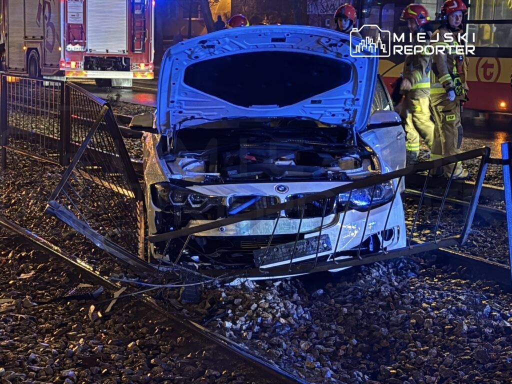 Uszkodzony samochód osobowy stoi na torach kolejowych, obok niego pracują strażacy. W tle widać woz strażacki oraz oświetlenie nocne.