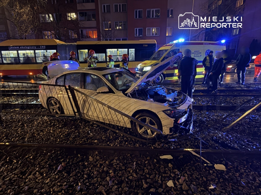 Biały samochód osobowy uszkodzony na torach tramwajowych, wokół strażacy i służby ratunkowe w akcji.