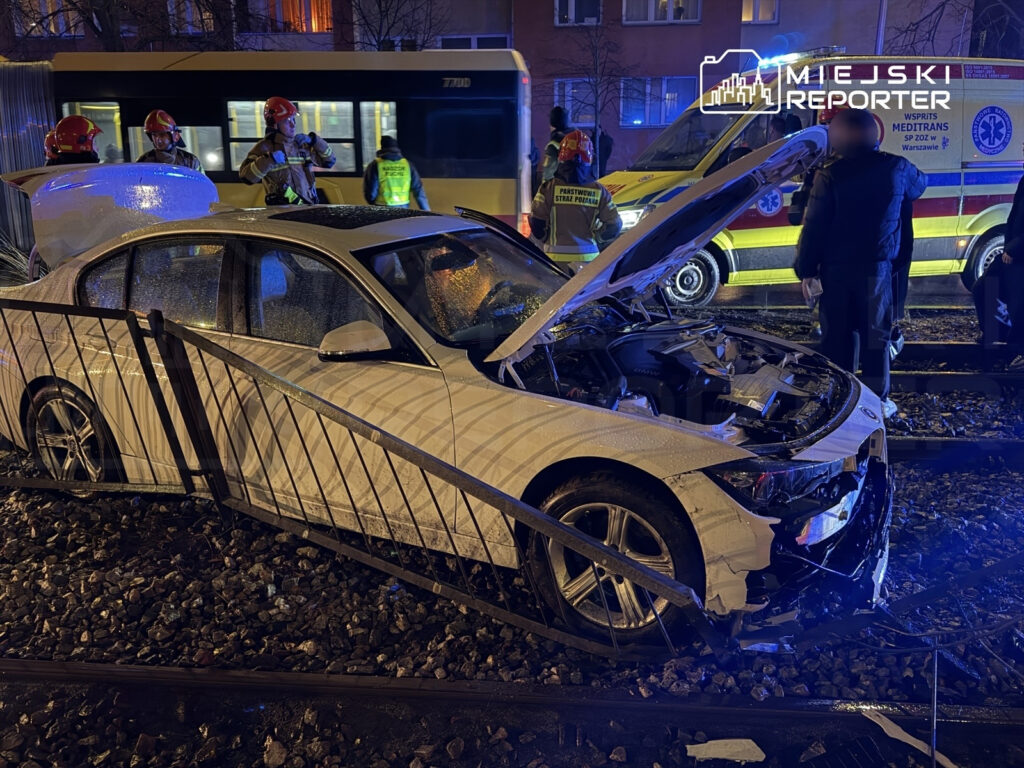 Biały samochód osobowy rozbity na torach, obok pracują strażacy i ratownicy medyczni. W tle widać autobus i karetki.