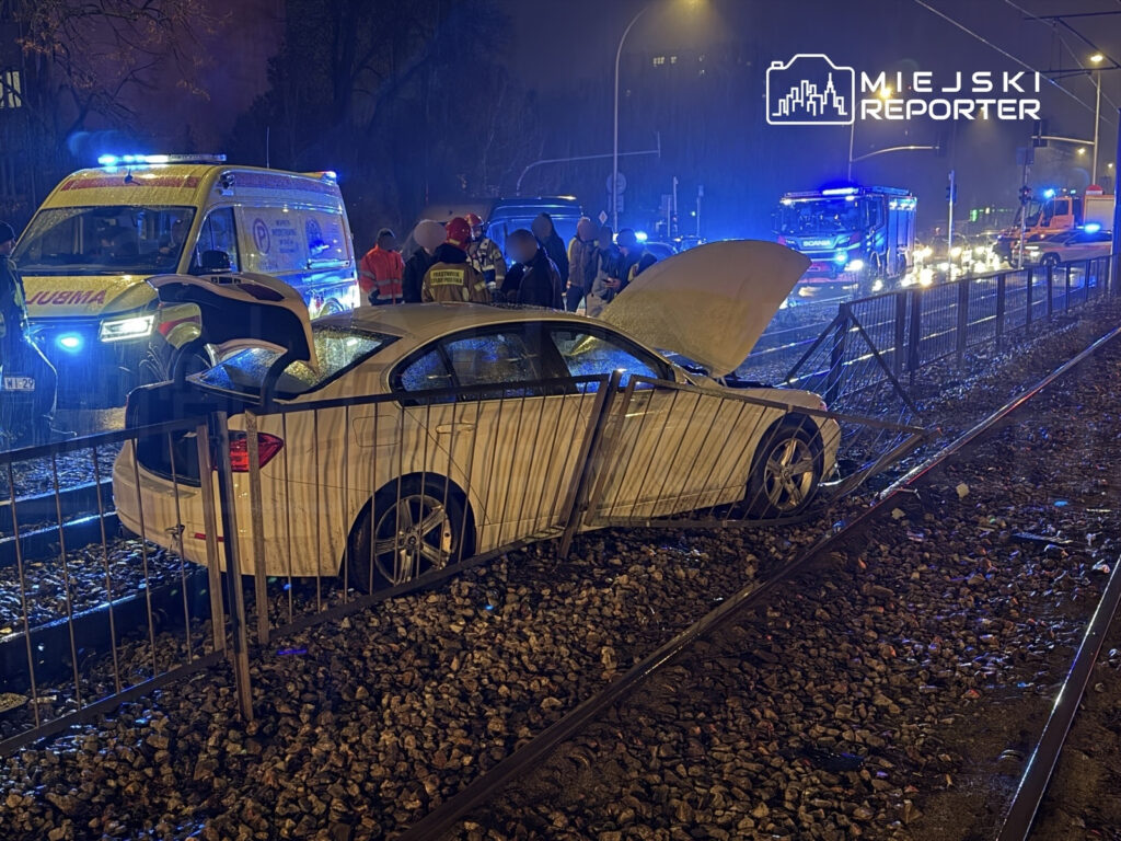 Biały samochód osobowy stoi na torach tramwajowych, otwarta pokrywa silnika, w tle działania służb ratunkowych.
