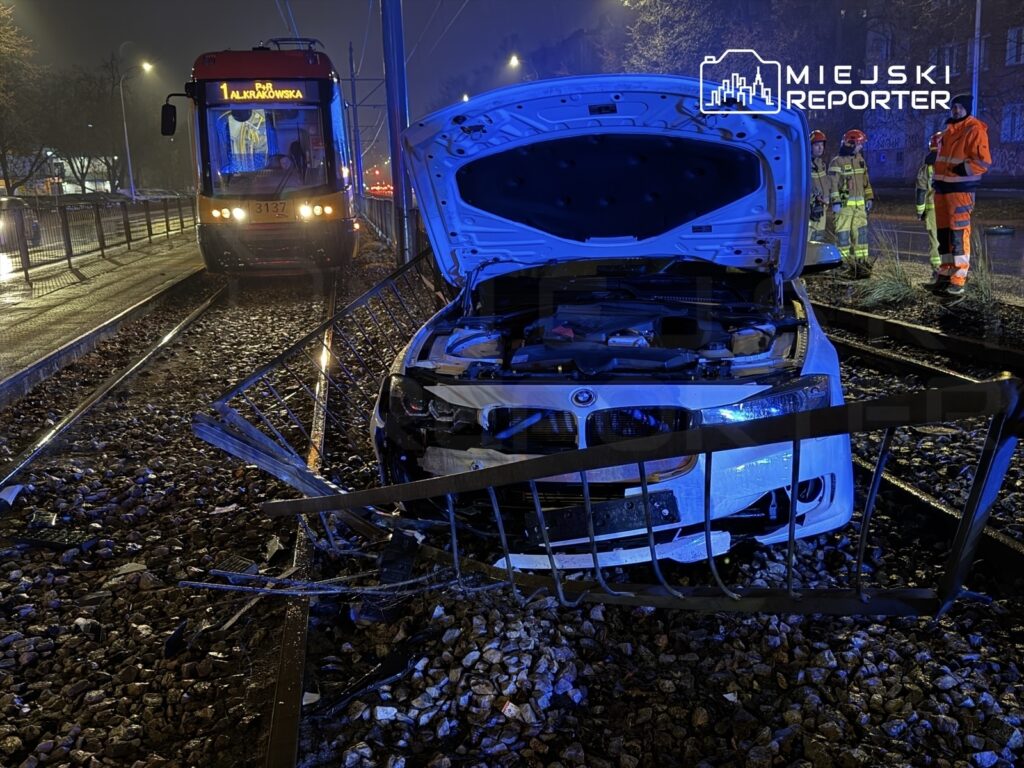 Biały samochód osobowy uszkodzony na torach tramwajowych, obok grupka strażaków w odblaskowych strojach.