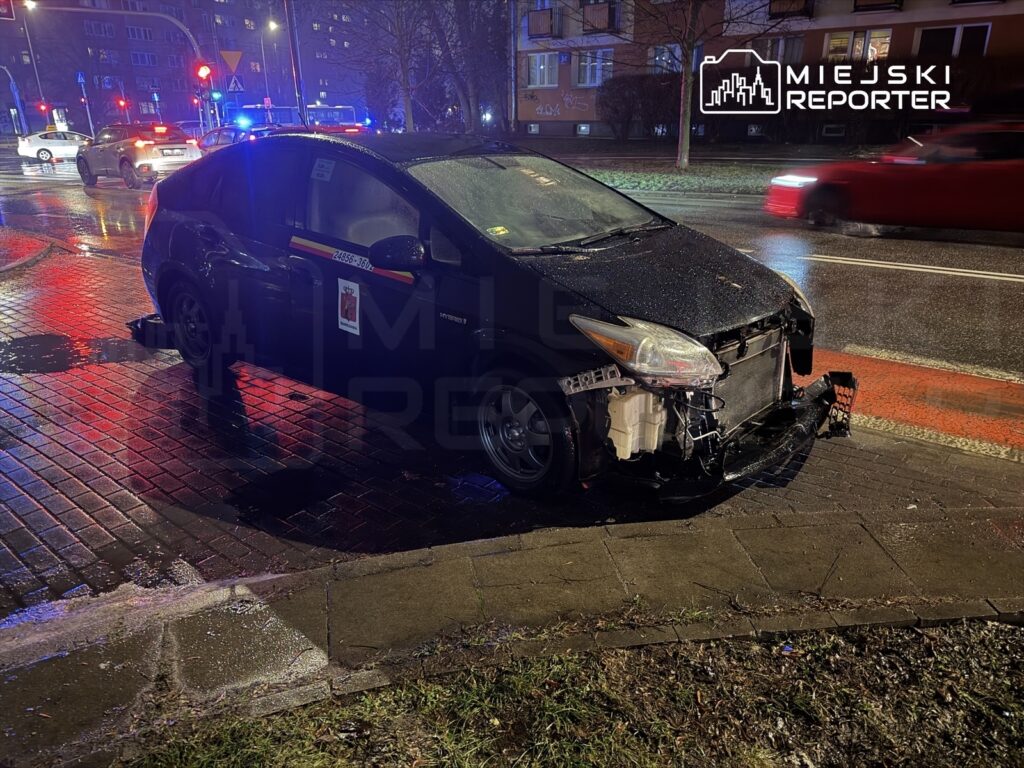Uszkodzony samochód osobowy stoi na chodniku w deszczową noc, obok widać migające światła policyjnych radiowozów.