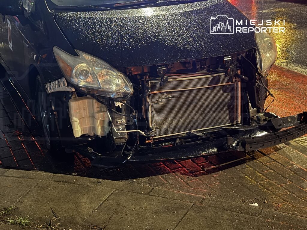 Uszkodzony samochód na ulicy z widoczną uszkodzoną przednią częścią, w tle mokra nawierzchnia po deszczu.