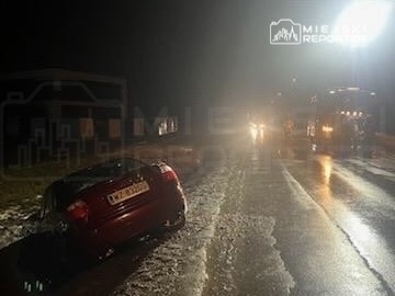 Czerwony samochód osobowy leży na boku przy drodze, w tle widać reflektory pojazdów ratunkowych w nocnym otoczeniu.