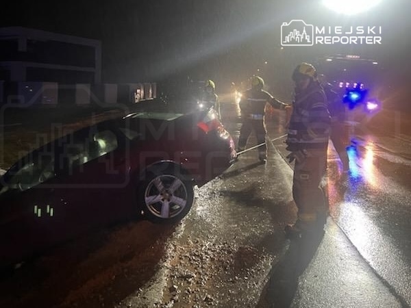 Czterech strażaków zabezpiecza miejsce zdarzenia, przy uszkodzonym samochodzie na mokrej drodze w nocy.