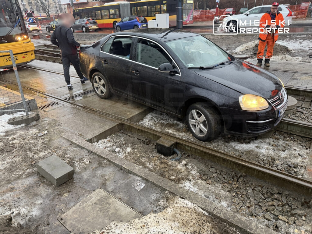 Czarny samochód osobowy utknął na torach tramwajowych, a mężczyzna w dresie rozmawia z kierowcą na ulicy.