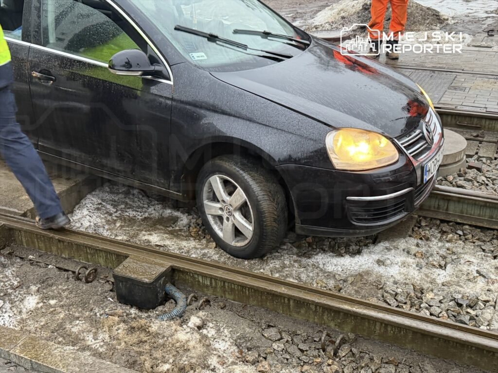 Czarny samochód osobowy stoi na torach kolejowych, obok mężczyzna w żółtej kamizelce roboczej.