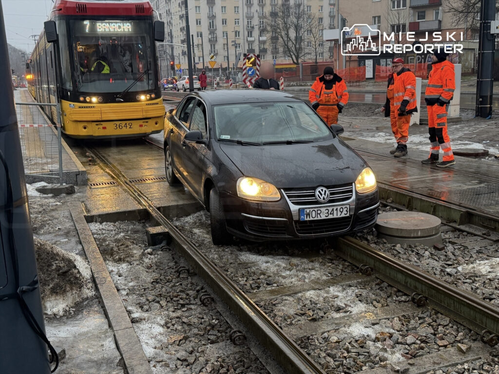 Czarny samochód osobowy utknął na torach tramwajowych, obok pracownicy w pomarańczowych ubraniach zabezpieczają miejsce.