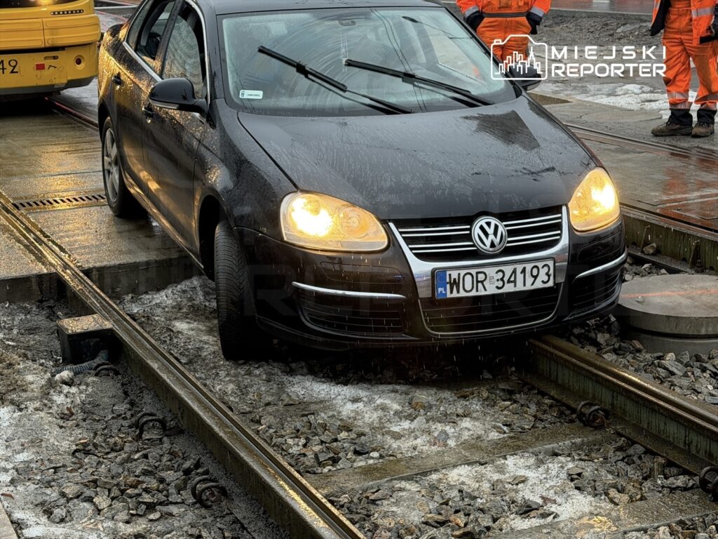 Czarny samochód osobowy stoi na torach kolejowych, wokół widoczne są pracownicy w pomarańczowych kombinezonach.