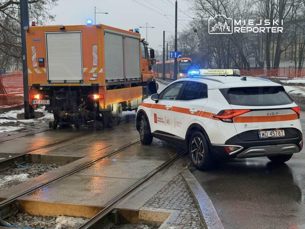 Pojazd służbowy z włączonymi sygnałami świetlnymi stoi na torowisku, obok drugiego pojazdu z pomarańczowym nadwoziem.