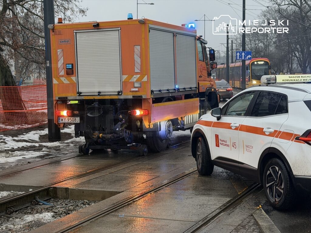 Żółty wóz strażacki z zamkniętą skrzynką i niebieskim światłem stoi na torach tramwajowych obok białego samochodu z pomarańczowymi detalami.