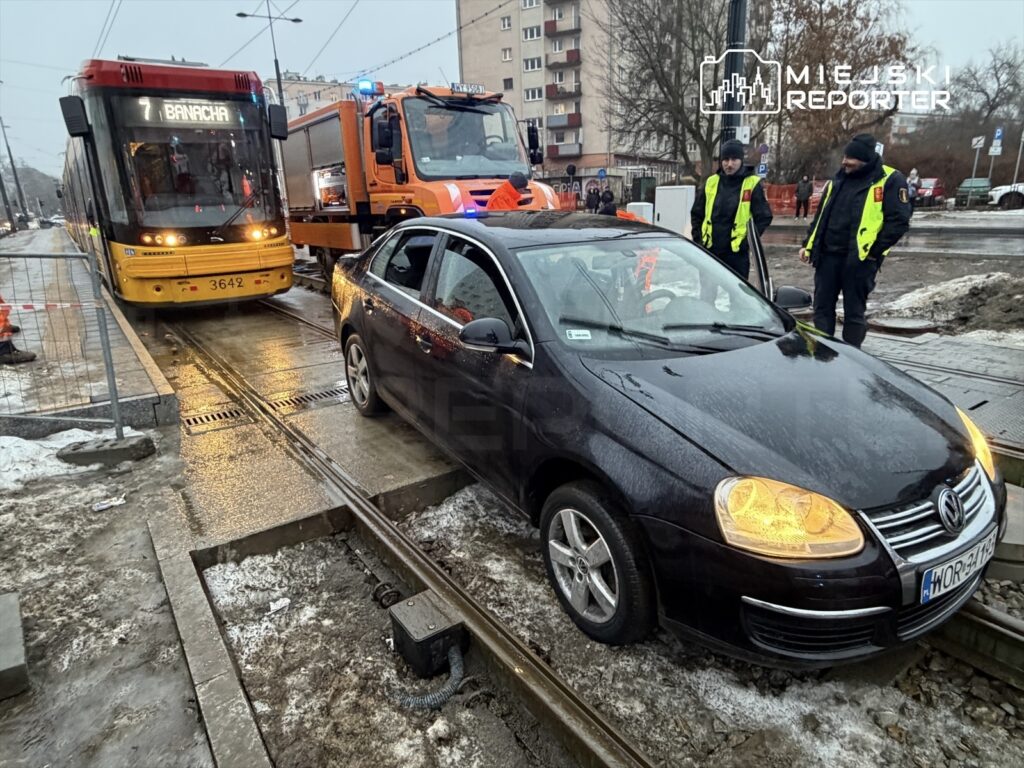 Czarny samochód osobowy utknął na torowisku tramwajowym, obok niego stoją funkcjonariusze w odblaskowych kurtkach.