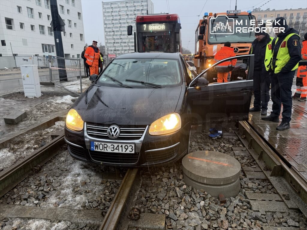 Czarny samochód osobowy stoi na torach tramwajowych, obok pracownicy w pomarańczowych i żółtych kamizelkach.