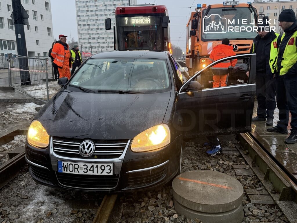 Czarny samochód osobowy stoi na torach tramwajowych, a obok grupka pracowników w pomarańczowych i żółtych kurtkach ocenia sytuację.