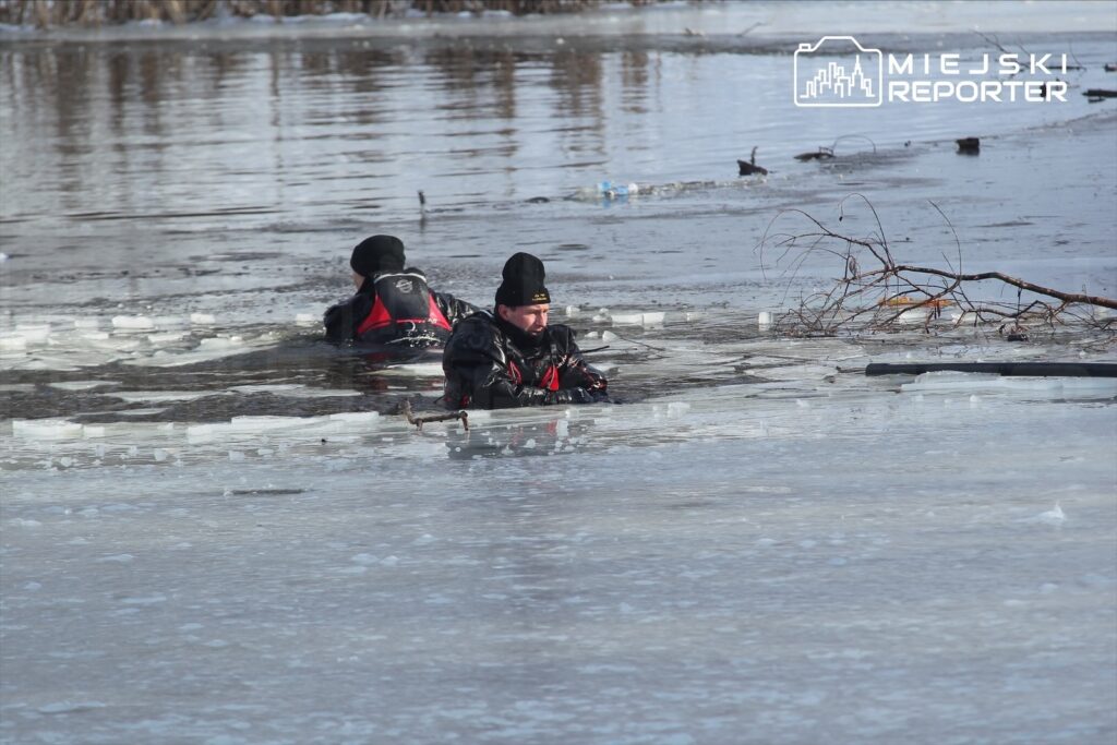 Dwóch mężczyzn w kombinezonach nurkowych przemieszcza się w wodzie pokrytej lodem.
