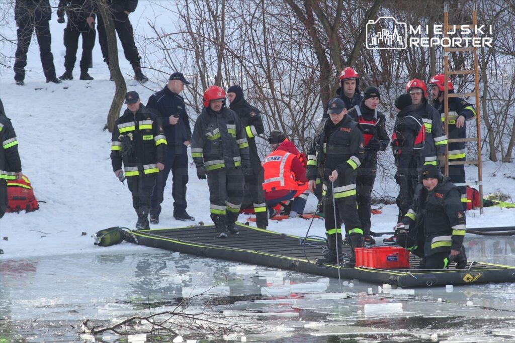 Grupa strażaków w specjalistycznych strojach pracuje na brzegu zamarzniętej rzeki, przygotowując sprzęt do akcji ratunkowej.