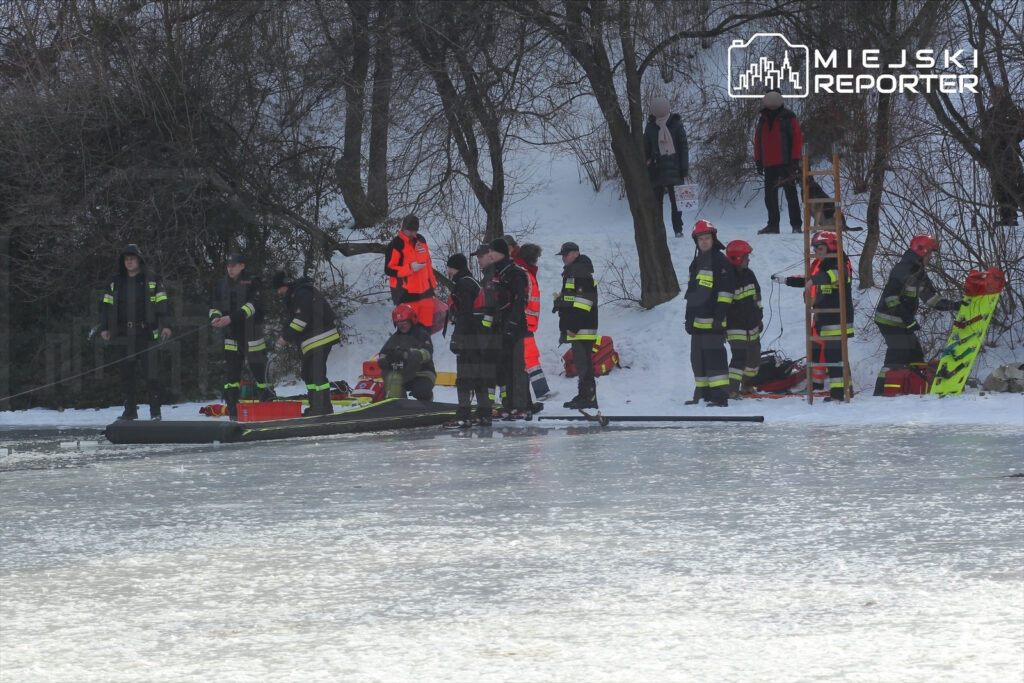 Grupa ratowników w strojach ochronnych przygotowuje sprzęt na brzegu zamarzniętego jeziora w zimowej scenerii.