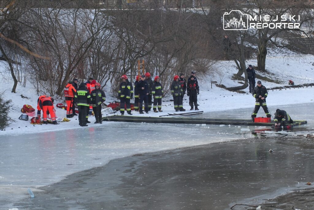 Grupa ratowników w strojach ochronnych przeprowadza akcję ratunkową na zamarzniętym jeziorze w zimowym otoczeniu.
