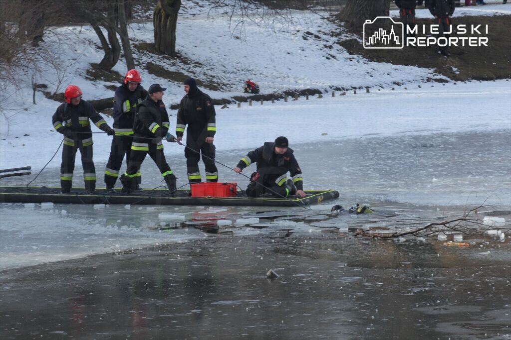 Grupa strażaków w specjalistycznych strojach na tratwie, pracująca nad ratowaniem osoby w zamarzniętym zbiorniku wodnym.