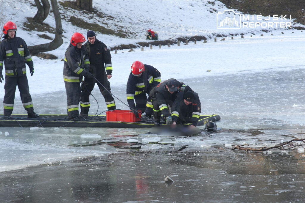 Grupa strażaków w specjalnych ubraniach ratuje osobę z zamarzniętego jeziora, otoczeni przez lód i śnieg.