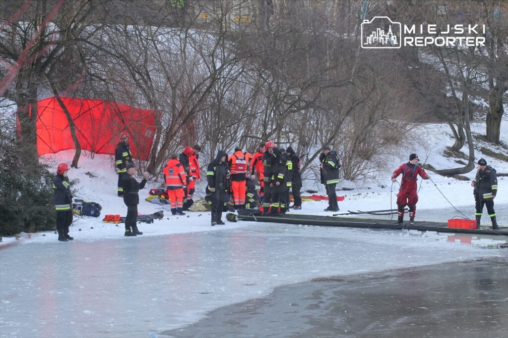 Grupa ratowników w specjalistycznych strojach pracuje nad zamarzniętym brzegiem zbiornika wodnego, w tle widoczna czerwona zasłona.