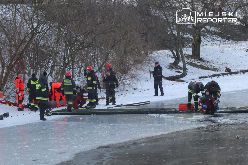 Grupa ratowników w specjalistycznych strojach pracuje nad osobą na zamarzniętej wodzie, w otoczeniu śniegu i drzew.
