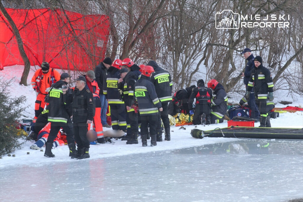 Grupa ratowników w strojach ochronnych udziela pomocy osobie leżącej na śniegu w pobliżu zamarzniętej wody.