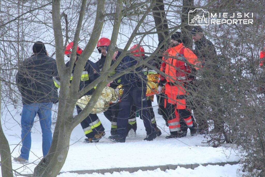 Grupa ratowników w mundurach niesie osobę owiniętą w folię termiczną przez zaśnieżony teren.
