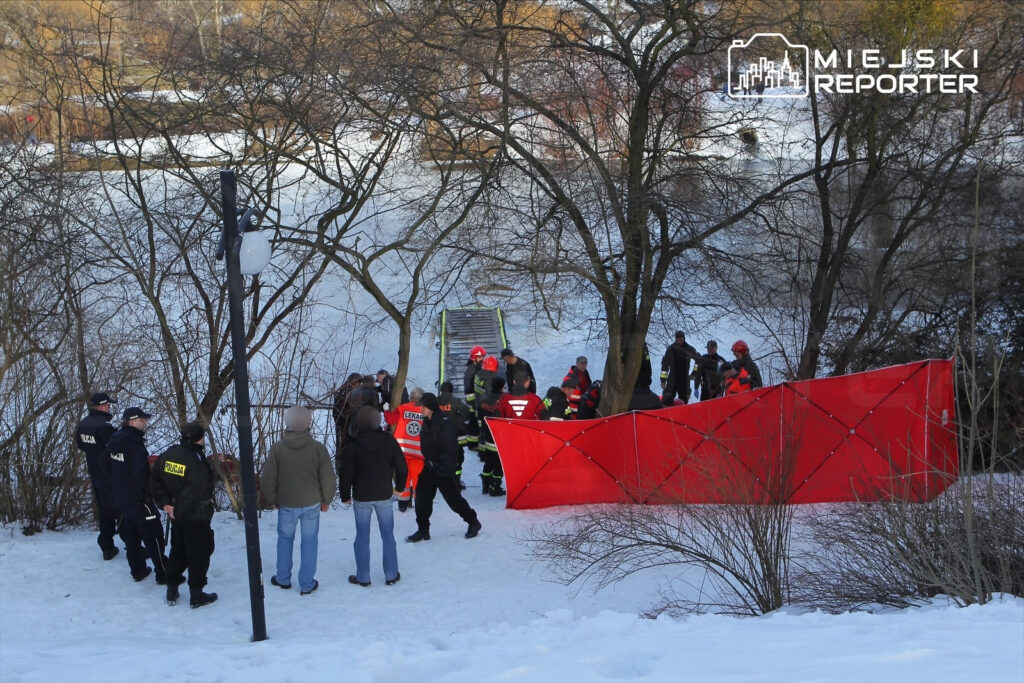 Grupa ratowników w odzieży ochronnej przeszukuje zamarznięty brzeg rzeki, otoczona przez policję i gapiów.