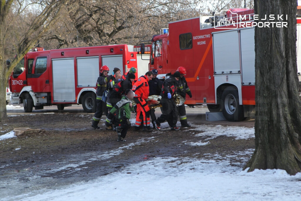 Grupa ratowników w mundurach niesie poszkodowanego w pobliżu wozu strażackiego na terenie parku.