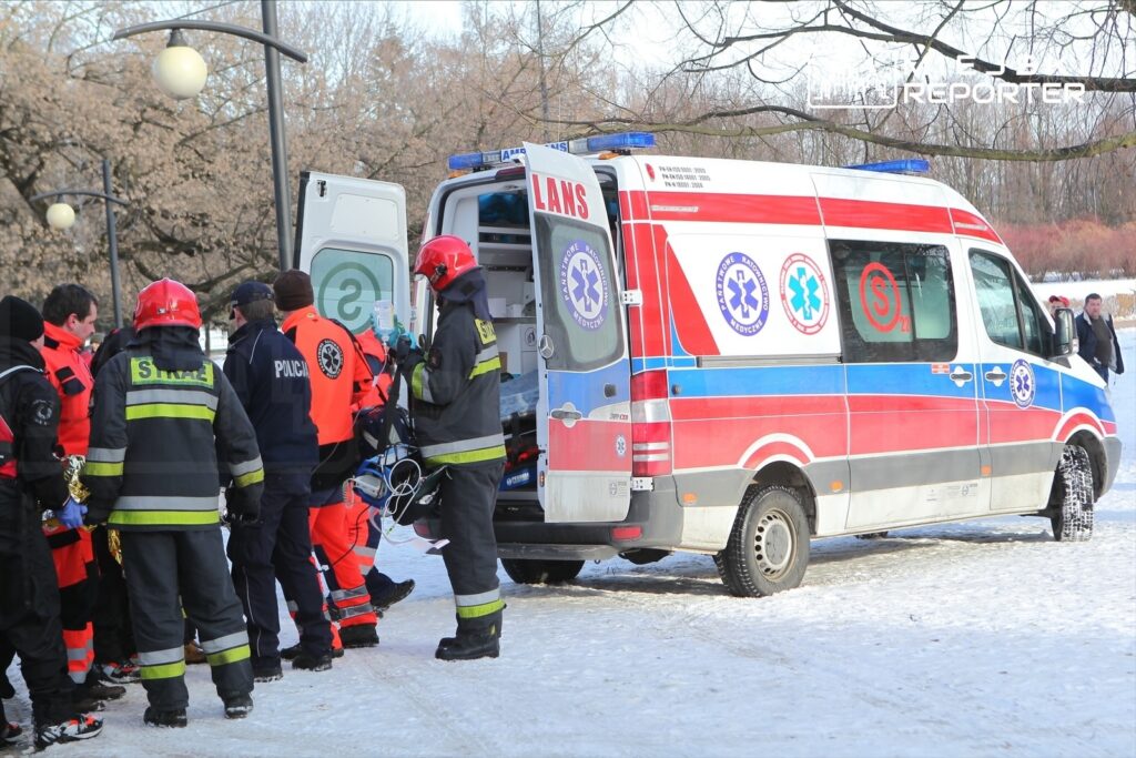 Zespół ratowników medycznych w akcji, otaczający ambulans na zaśnieżonym terenie.