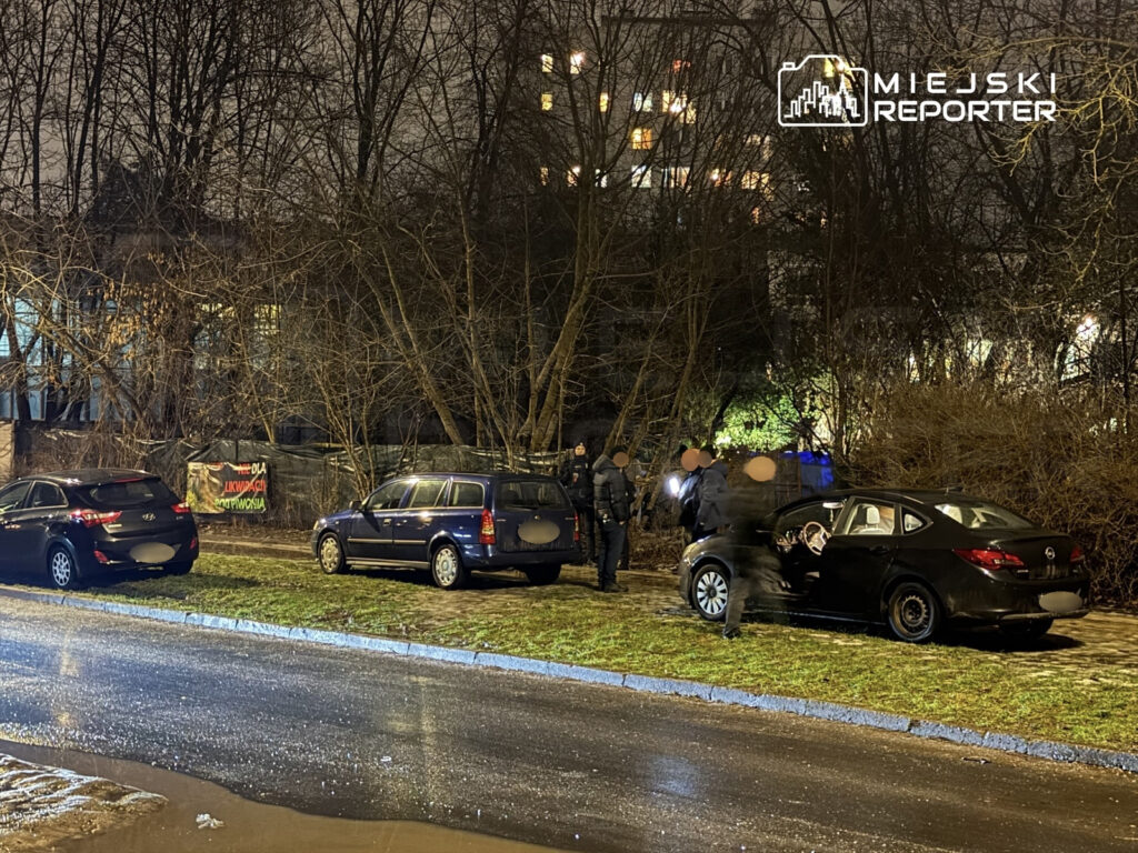 Grupa osób w odzieży roboczej przeszukuje samochód na tle nocy, obok zaparkowane pojazdy i nagranie z wydarzenia.