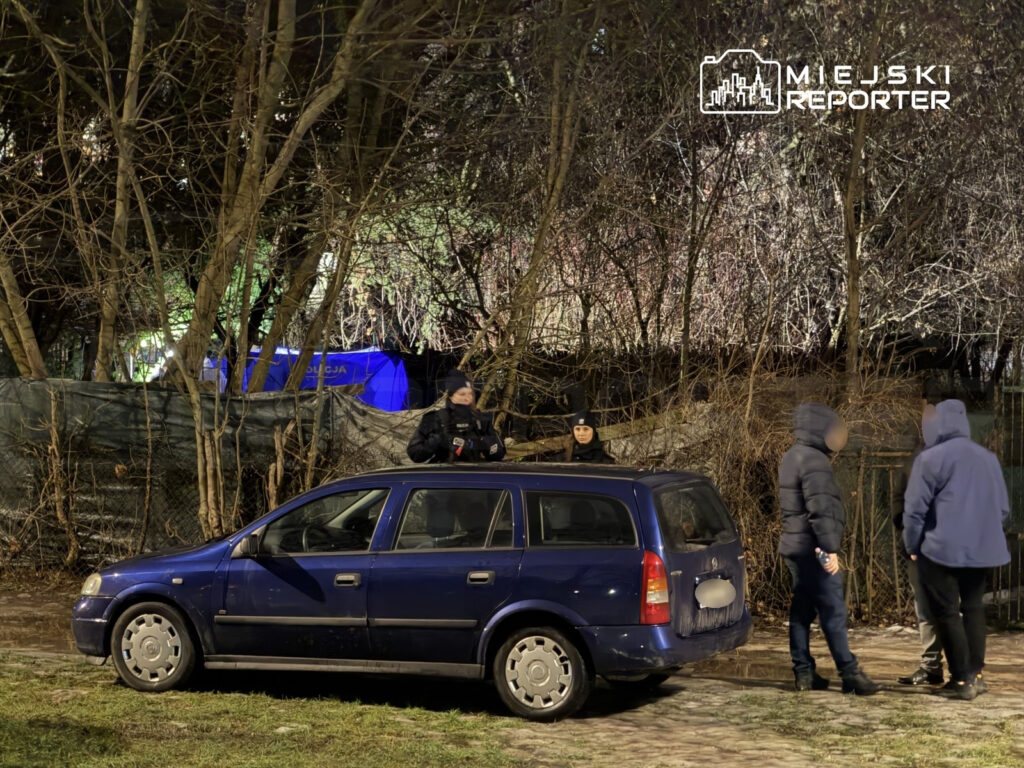 Grupa osób w ciemnych ubraniach rozmawia obok niebieskiego samochodu na oświetlonym terenie z drzewami w tle.