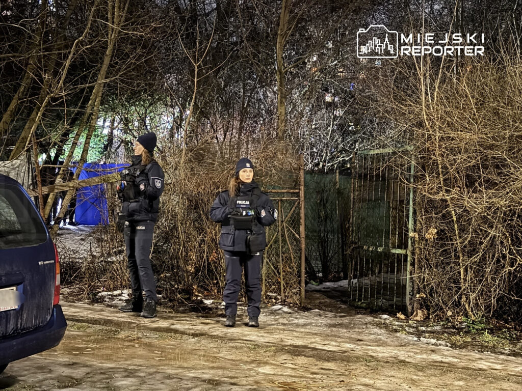 Dwóch policjantów stoi na zewnątrz, w otoczeniu zarośli, jeden z nich trzyma broń, a w tle widać niebieski namiot.