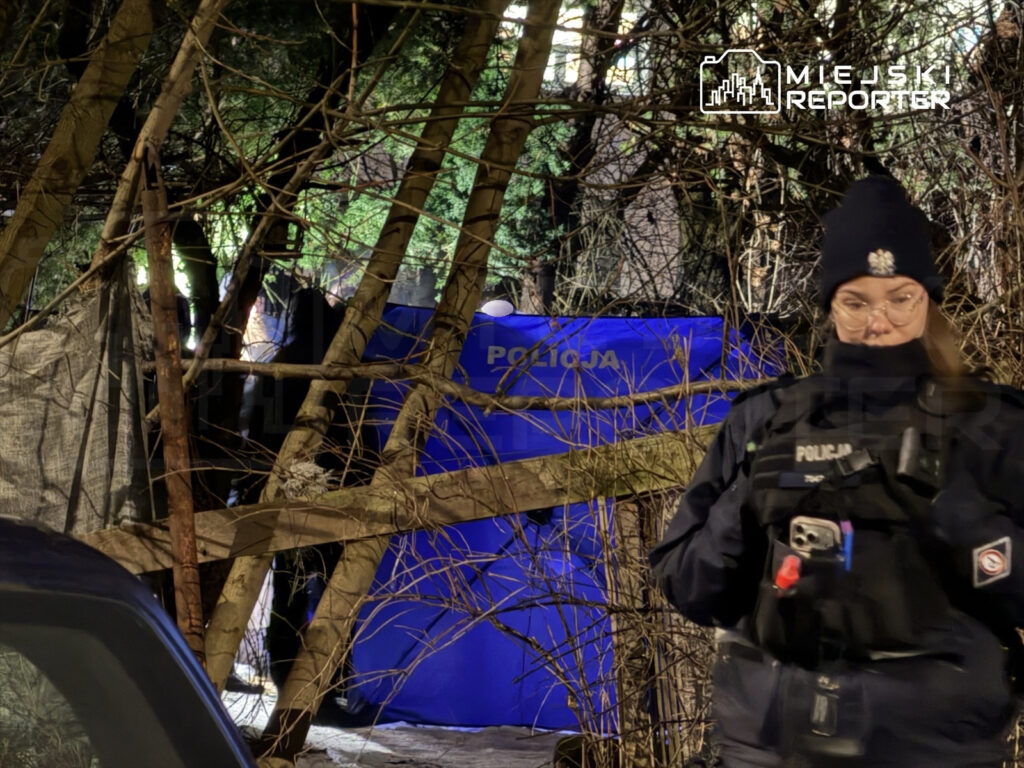 Policjantka w mundurze stoi w pobliżu niebieskiego namiotu z napisem "POLICJA", otoczona drzewami i gałęziami.