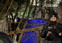 Policjantka w mundurze stoi w pobliżu niebieskiego namiotu z napisem "POLICJA", otoczona drzewami i gałęziami.