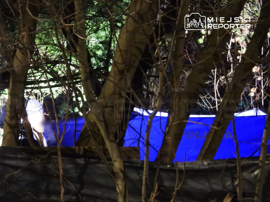 Mężczyzna w ciemnej odzieży przebywa w pobliżu niebieskiego namiotu policyjnego otoczonego drzewami.