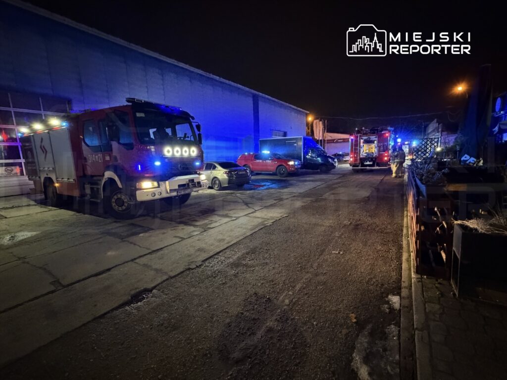 Czerwony wóz strażacki z włączonymi sygnałami świetlnymi stoi na oświetlonym podjeździe w nocy, obok innych pojazdów.